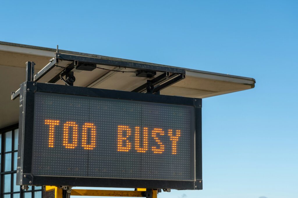 Eine digitale LED-Anzeigetafel, ähnlich einem Mautstation- oder Baustellenhinweis, zeigt in orangenen Lettern die Warnung ‚Too Busy‘ und symbolisiert den Zustand hoher Auslastung ohne erkennbaren Fortschritt.
