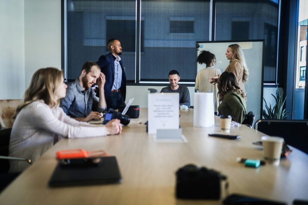 Mehrere Teilnehmende sitzen in einem unübersichtlichen Meeting zusammen; einige sprechen gleichzeitig, während andere abgelenkt oder ratlos wirken.