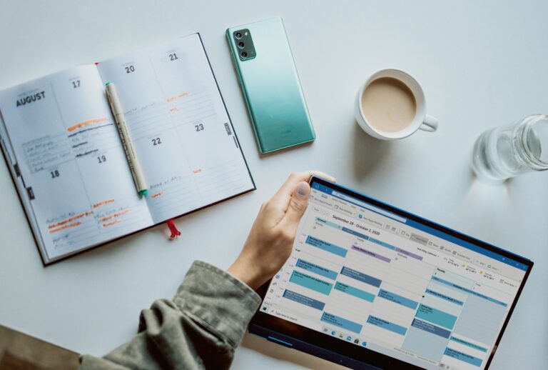 Vogelperspektive auf einen Arbeitstisch mit einem aufgeklappten Kalender oder Planer auf der linken Seite. Rechts hält eine Hand ein Tablet mit einem dicht gefüllten Microsoft-Outlook-Kalender.