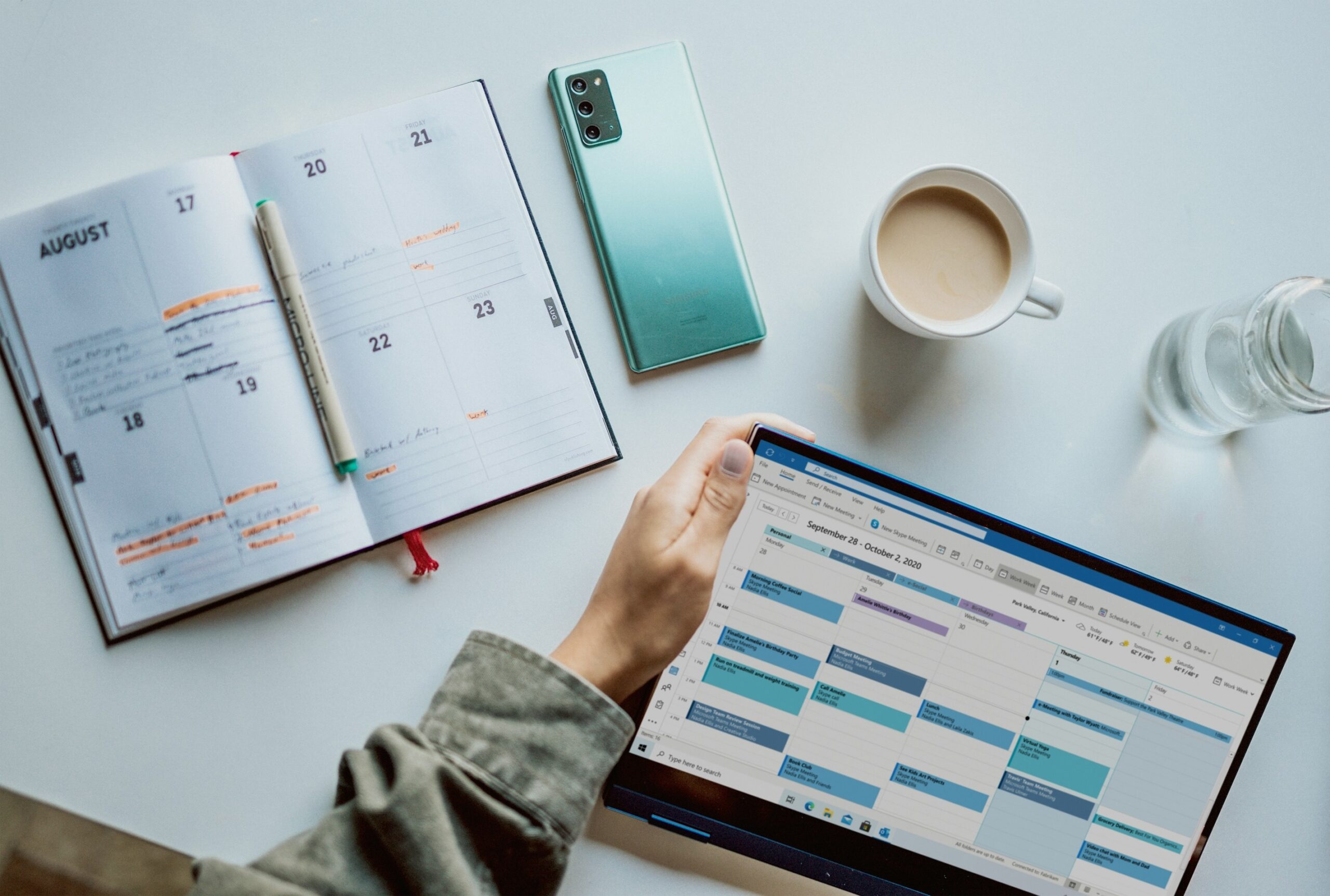 Vogelperspektive auf einen Arbeitstisch mit einem aufgeklappten Kalender oder Planer auf der linken Seite. Rechts hält eine Hand ein Tablet mit einem dicht gefüllten Microsoft-Outlook-Kalender.